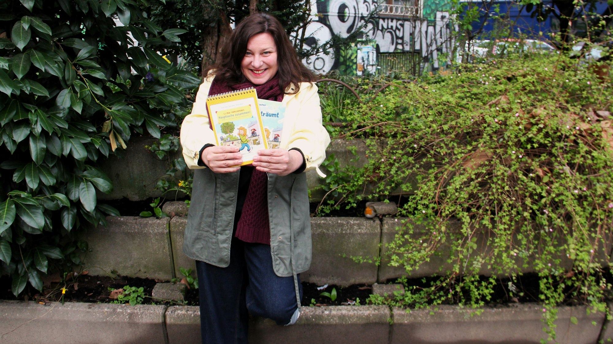 Eine Frau mit dunklem steht vor einem Stufen-Beet und zeigt zwei Kinderbücher.