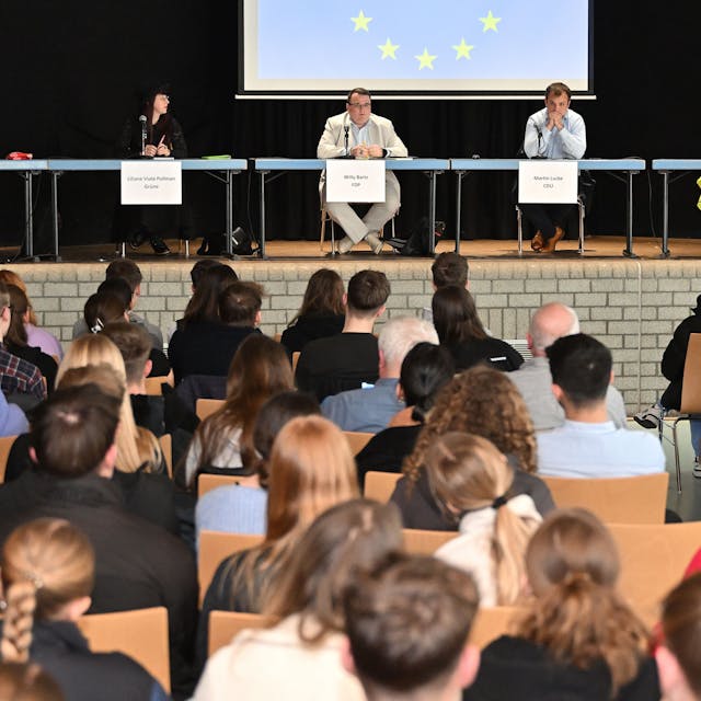 Jugendliche sitzen im Publikum, auf der Bühne sitzen Politiker mit Namensschildern. Hinter ihnen die Europafahne.