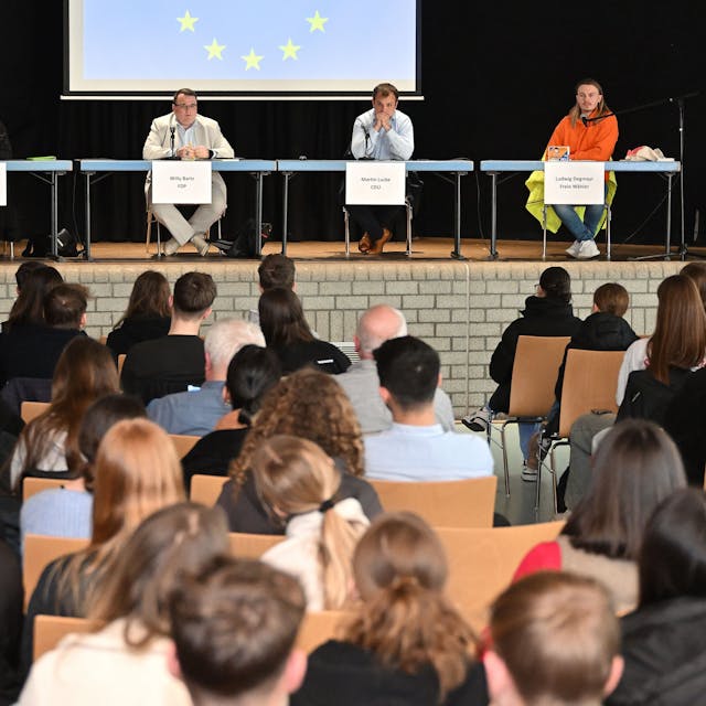 Jugendliche sitzen im Publikum, auf der Bühne sitzen Politiker mit Namensschildern. Hinter ihnen die Europafahne.