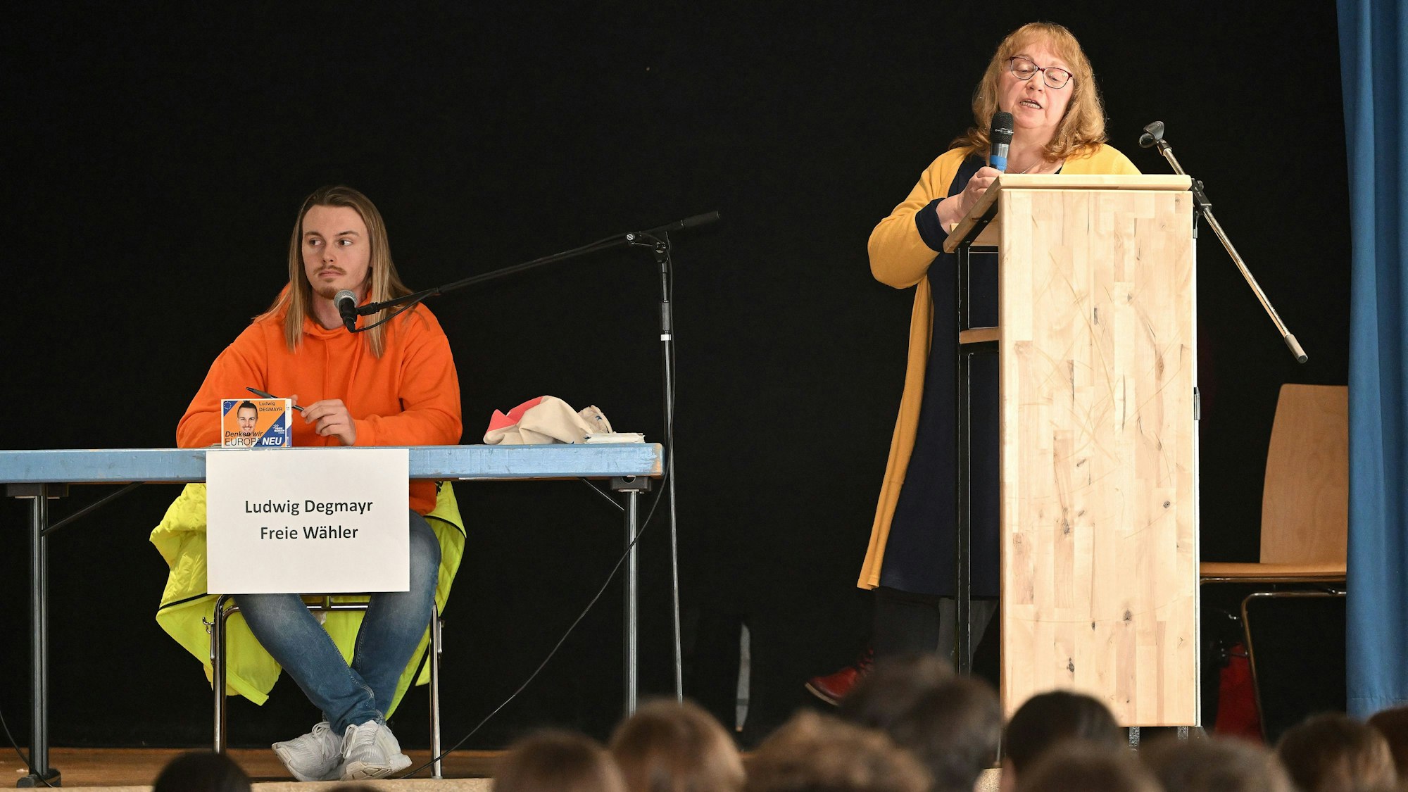 Ein junger Politiker sitzt auf dem Podium, eine Frau steht am Rednerpult und spricht in ein Mikrofon.