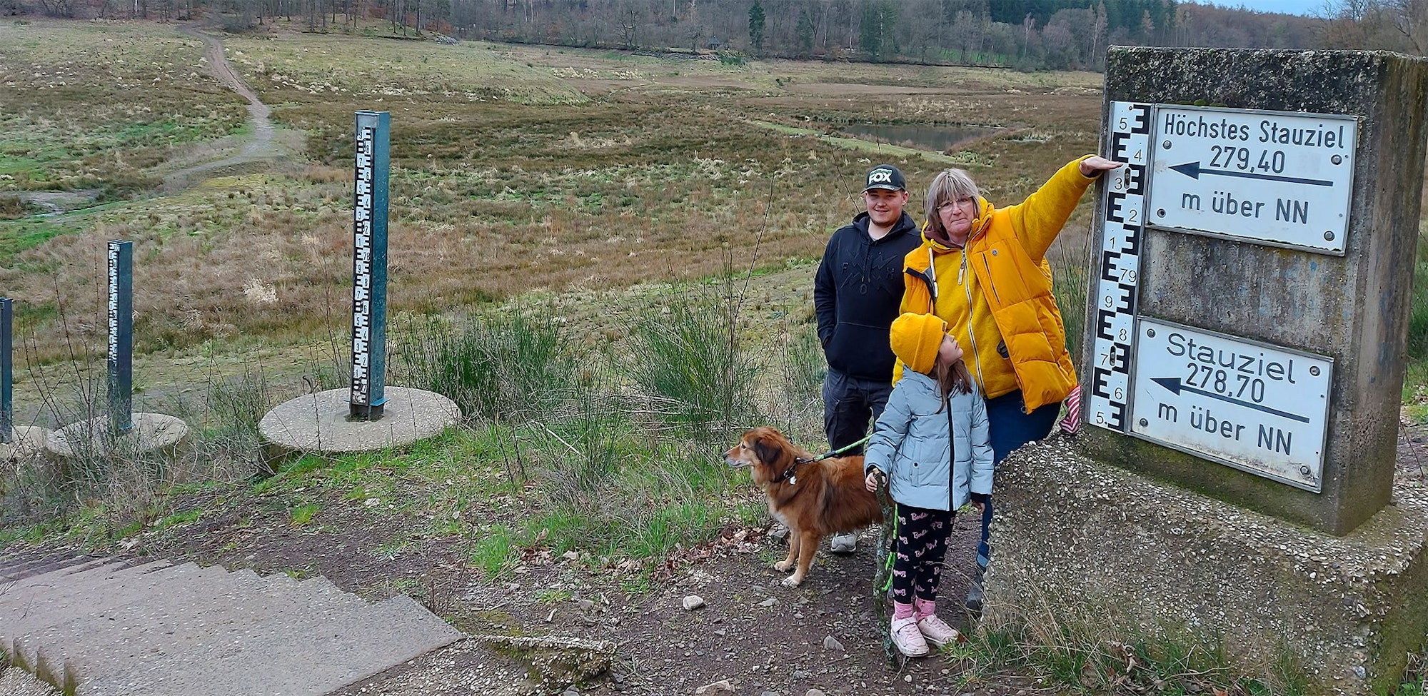 Zwei Erwachsene, ein Kind und ein Hund stehen vor den Pegel-Anzeigen. Die Frau zeigt auf ein Schild mit der Aufschrift „Höchstes Stauziel 279,40 m über NN“.