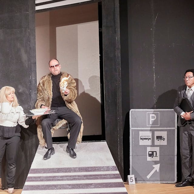 Eine Frau und zwei Männer, einer im Pelzmantel, der andere im Anzug, stehen auf der Bühne.