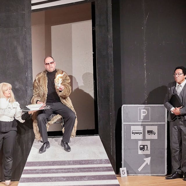 Eine Frau und zwei Männer, einer im Pelzmantel, der andere im Anzug, stehen auf der Bühne.