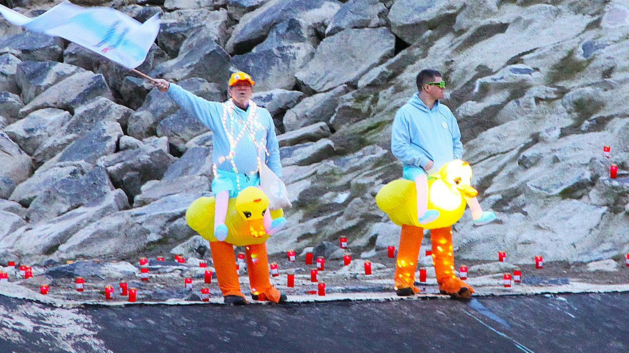 Zwei Männer, die gelbe Entchen-Kostüme umgebunden haben, stehen an der Steinbach. Einer schwenkt eine hellblaue Fahne.