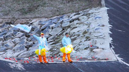 Zwei Männer, die gelbe Entchen-Kostüme umgebunden haben, stehen an der Steinbach. Einer schwenkt eine hellblaue Fahne.