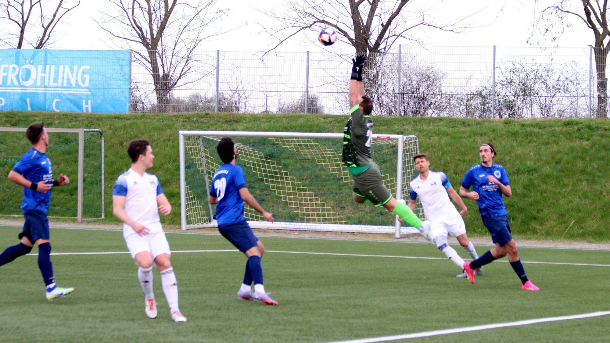 D-H-O-Torwart Dominik Schöpfer springt bei voller Körperspannung in die Luft. Der Ball scheint unerreichbar. Mit- und Gegenspieler schauen zu.