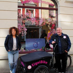 Eine Frau mit gelocktem Haar und ein Mann mit grauem Haar stehen neben einem Lastenrad mit der Aufschrift EigelBike.