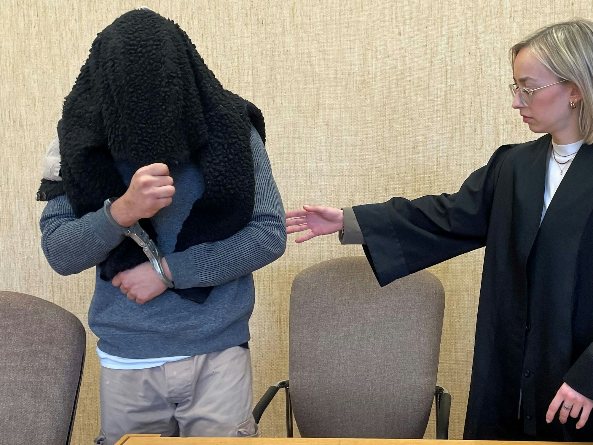 Ein Mann in Handschellen und mit einem Pullover über Kopf und Gesicht nimmt auf der Anklagebank Platz. Seine Anwältin leitet ihn mit der Hand.