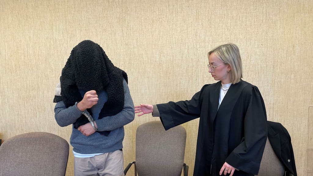 Ein Mann in Handschellen und mit einem Pullover über Kopf und Gesicht nimmt auf der Anklagebank Platz. Seine Anwältin leitet ihn mit der Hand.