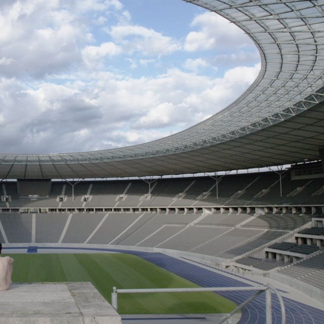 Das Bild zeigt, wie Eszter Salamon auf einem Plateau sitzt und auf ein riesiges Stadion im Hintergrund schaut.&nbsp;