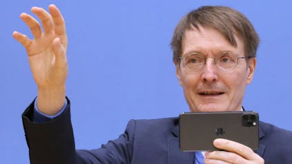 Karl Lauterbach hält sein Handy in der linken Hand und gestikuliert mit der rechten. Er trägt einen dunkelblauen Anzug. Der Hintergrund ist ebenfalls blau.&nbsp;