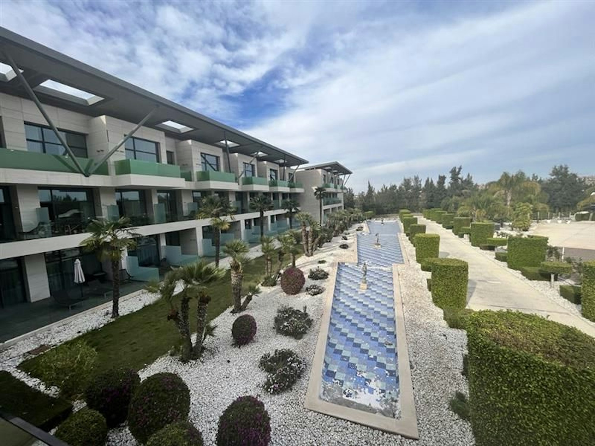 Blick auf die Hotelanlage von Innen. In der Mitte ein langes Wasserbecken. Links davon Palmen, rechts grüne Büsche.