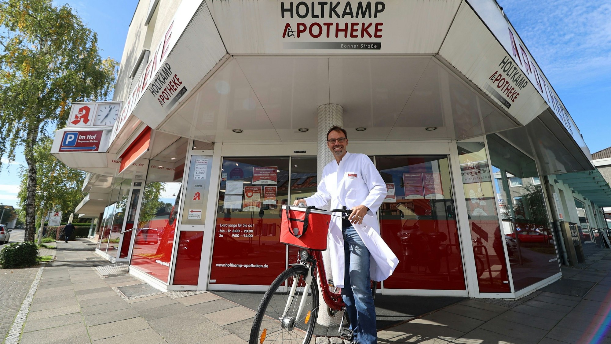 Ein Apotheker im weißen Arztkittel vor der Ladentür. Er steht neben einem roten Fahrrad.