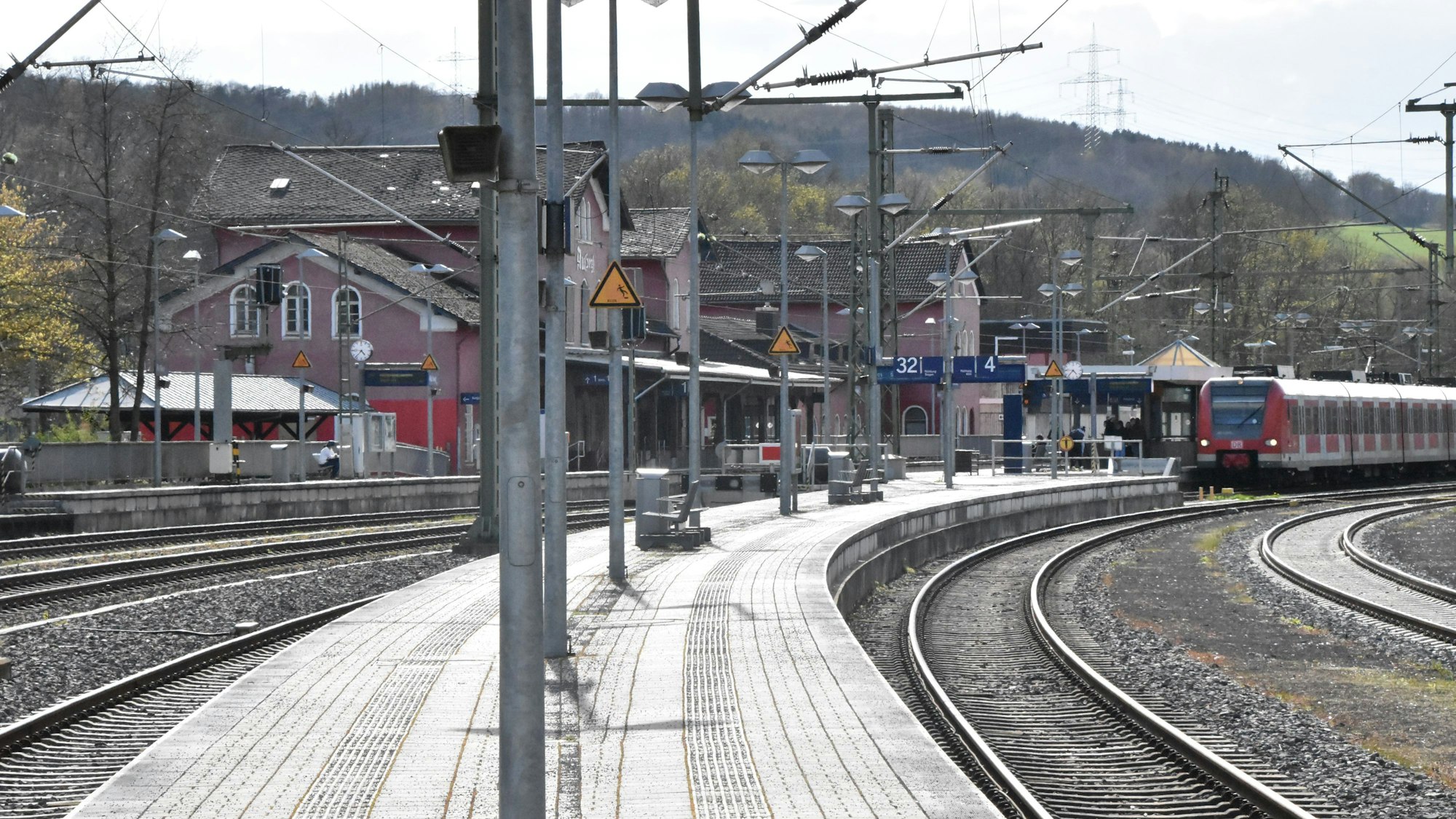 Ein Zug fährt in den Bahnhof ein, dessen rötliches Gebäude hinter gebogenen Bahnsteigen liegt. Gleise und Bahnsteige sind sauber.