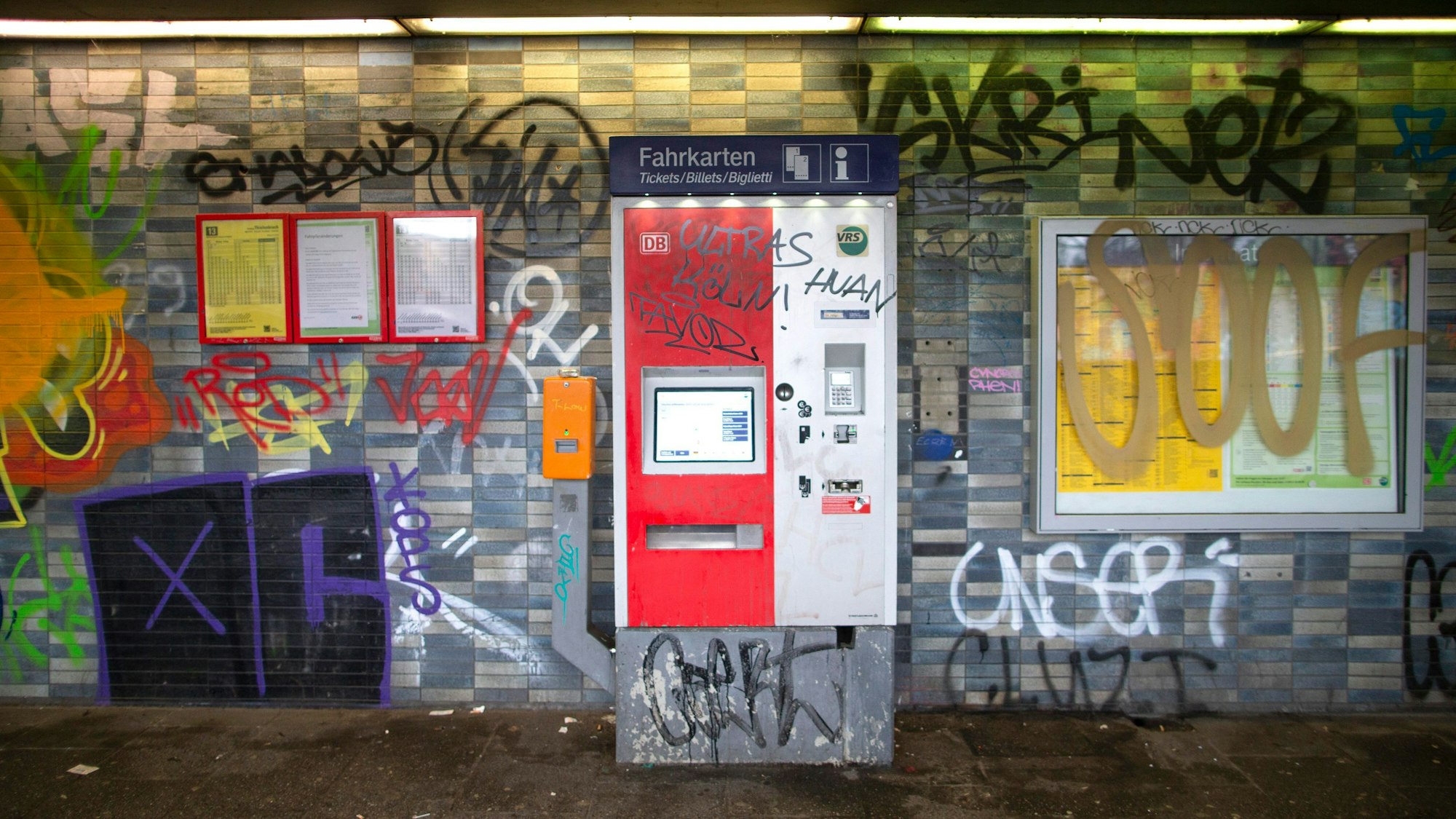 Eine gekachelte Wand in einem Bahnhof sowie ein Fahrkartenautomat und Fahrplanaushänge sind mit Graffiti und Schriftzügen beschmiert.