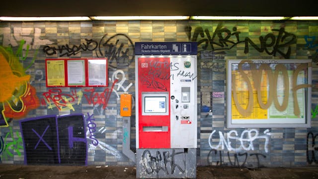Eine gekachelte Wand in einem Bahnhof sowie ein Fahrkartenautomat und Fahrplanaushänge sind mit Graffiti und Schriftzügen beschmiert.&nbsp;