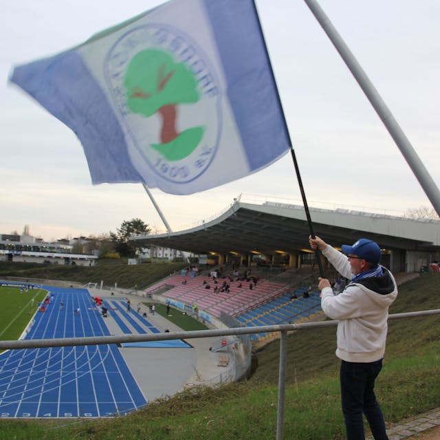Ein Fan schwenkt eine große Fahne des TuS Blau-Weiß Königsdorf. Im Hintergrund ist die Haupttribüne des Bonner Stadions zu sehen.