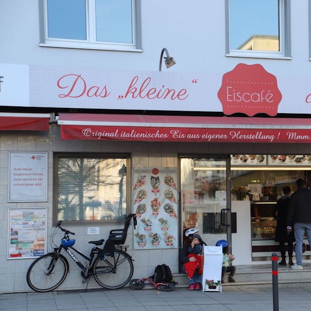 Das kleine Eiscafe am Markt in Eitorf in der Außenansicht. Menschen bestellen an der Eistheke, links im Bild ist ein Fahrrad mit Kindersitz an die Hauswand gelehnt.