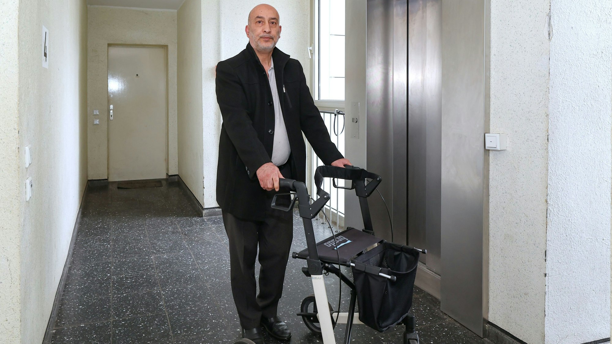 Zu sehen ist Halis Yildirim aus Bocklemünd in der Vorderansicht vor einem Aufzug mit Rollator stehend.
