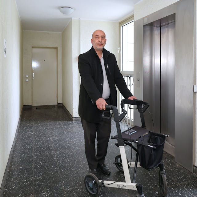 Zu sehen ist Halis Yildirim aus Bocklemünd in der Vorderansicht vor einem Aufzug mit Rollator stehend.
