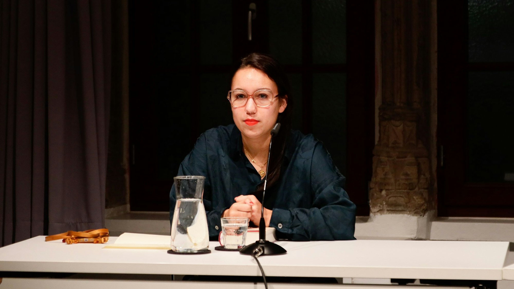 Ronya Othmann sitzt an einem Tisch, vor ihr ist ein Mikrofon aufgestellt. Auf dem Tisch stehen auch ein Glas und eine Karaffe mit Wasser. Die Autorin hat dunkle Haare, trägt eine Brille und eine dunkelgrüne Bluse.