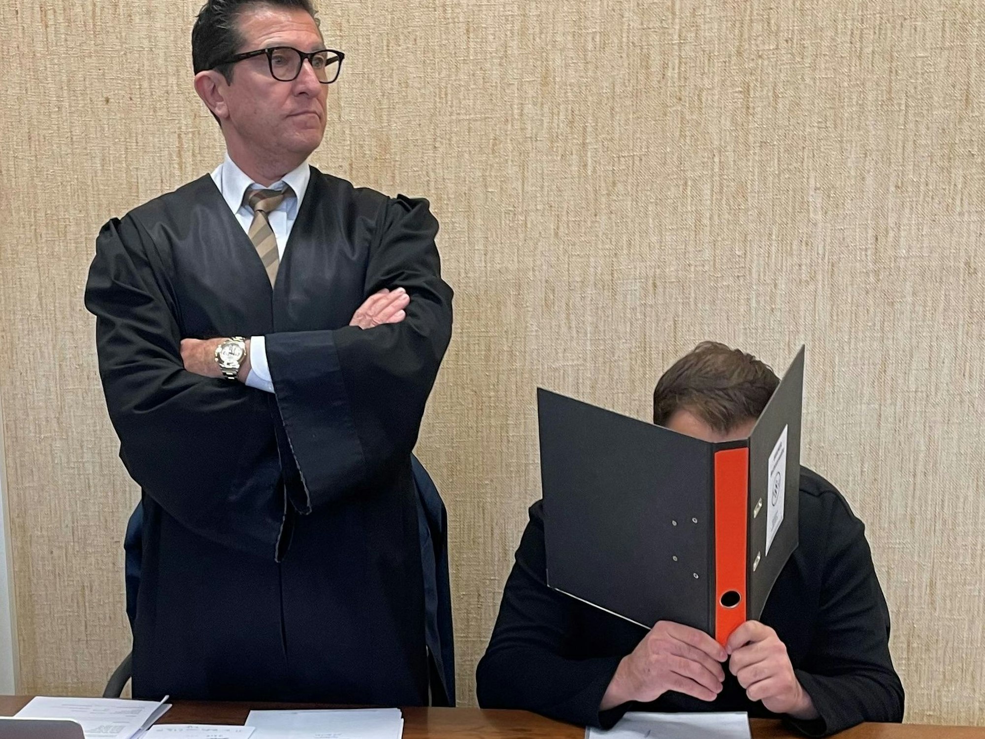 Ein Mann sitzt auf der Anklagebank und hält sich einen Aktenordner vor das Gesicht, neben ihm steht sein Anwalt.