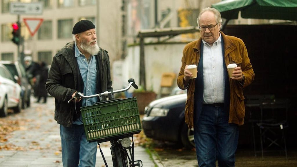 Zwei ältere Männer gehen während einer TV-Szene mit einem Fahrrad an einer Straße entlang.