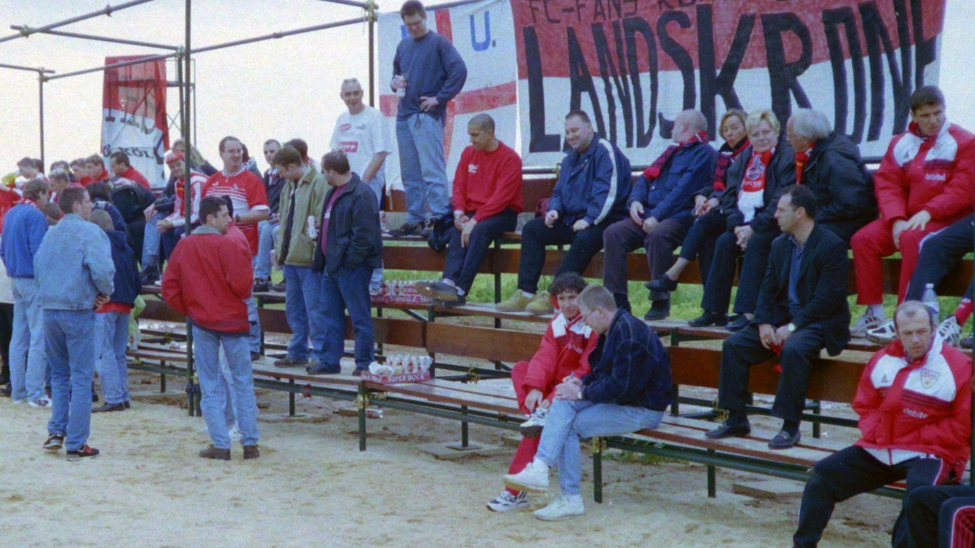 Mehrere Fußball-Fans teilen sich eine kleine Tribüne mit Funktionären des gegnerischen Vereins.