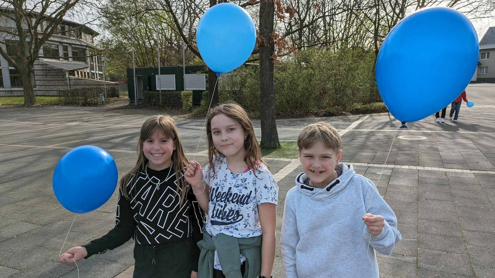 Drei Schüler und Schülerinnen stehen mit je einem Ballon in der Hand auf dem Schulhof.