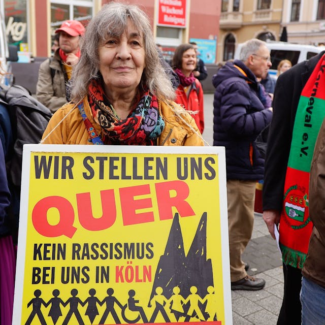 Menschen bei einer Demonstration. Eine Frau trägt ein Plakat mit der Aufschrift: „Wir stellen quer - Kein Rassismus bei uns in Köln“.&nbsp;