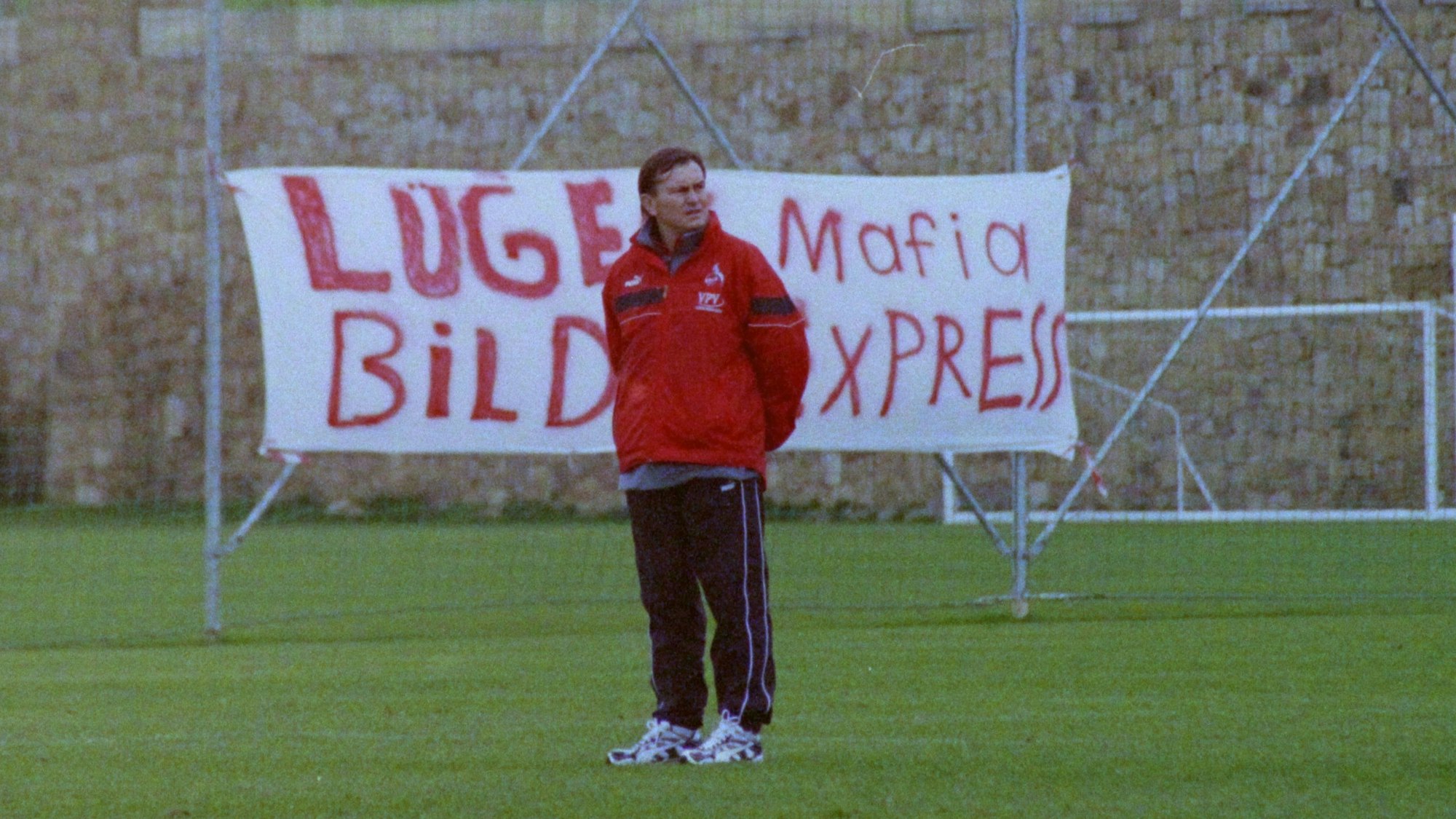 Ein Fußball-Trainer steht auf einem Sportplatz und beobachtet seine Mannschaft. Im Hintergrund ist ein Protestplakat zu erkennen.