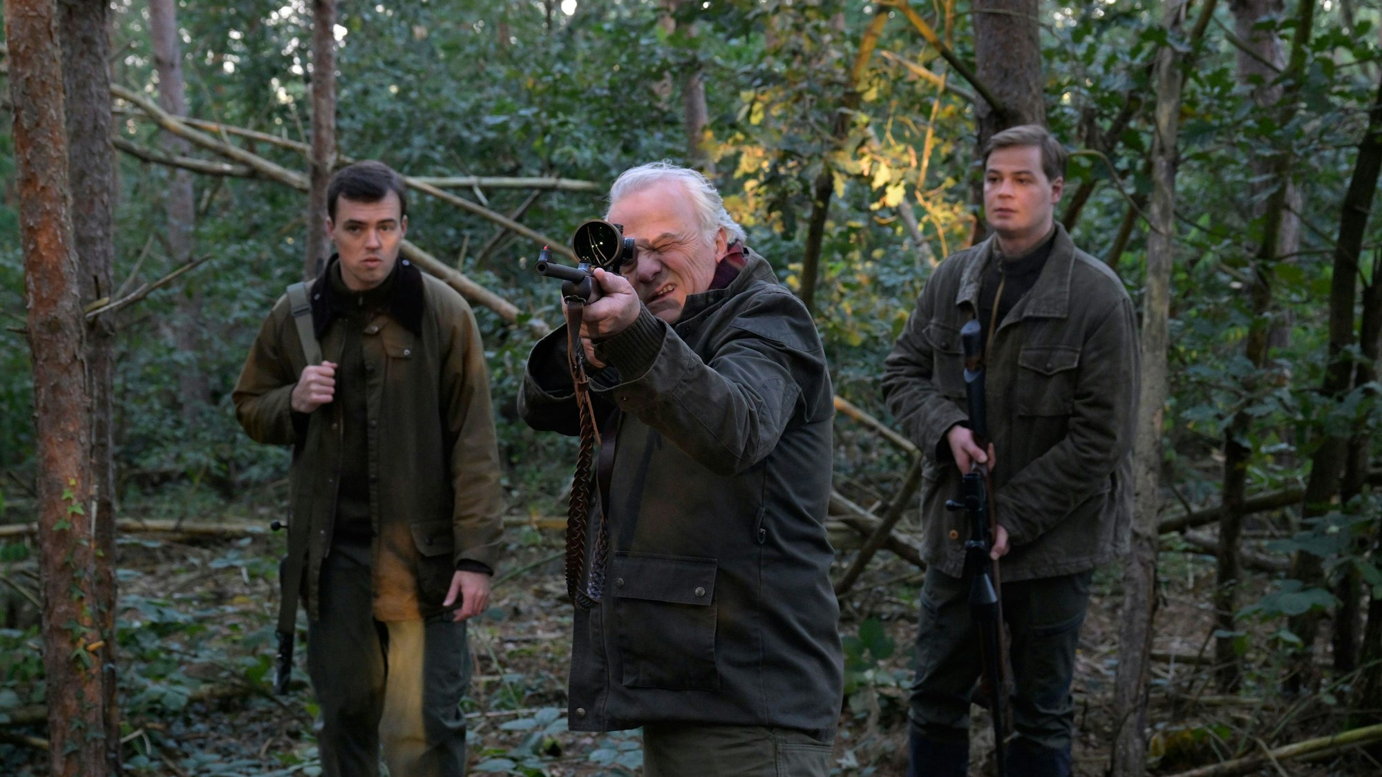 Die drei Charaktere stehen im Wald. Dr. Albrecht Richtmann, der Chef der Kanzlei, zielt mit einem Jagdgewehr. Er steht mittig zwischen seinem Sohn Konstantin Richtmann und Daniel Pillokat, die beide der gleichen Anwaltskanzlei des Schützen arbeiten.