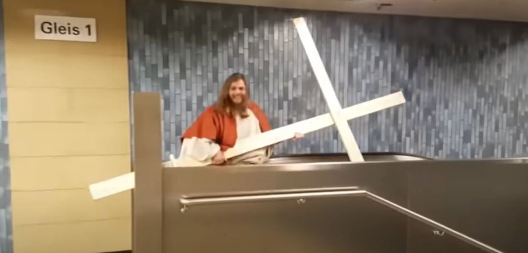 Ausschnitt aus einem Youtube-Video, das einen "Jesus" mit Holzkreuz auf der Rolltreppe in der KVB-Haltestelle Dom/Hauptbahnhof zeigt. In der Decke sind zwei Löcher zu erkennen, die er mit dem Holzkreuz dort hinterlassen hat.