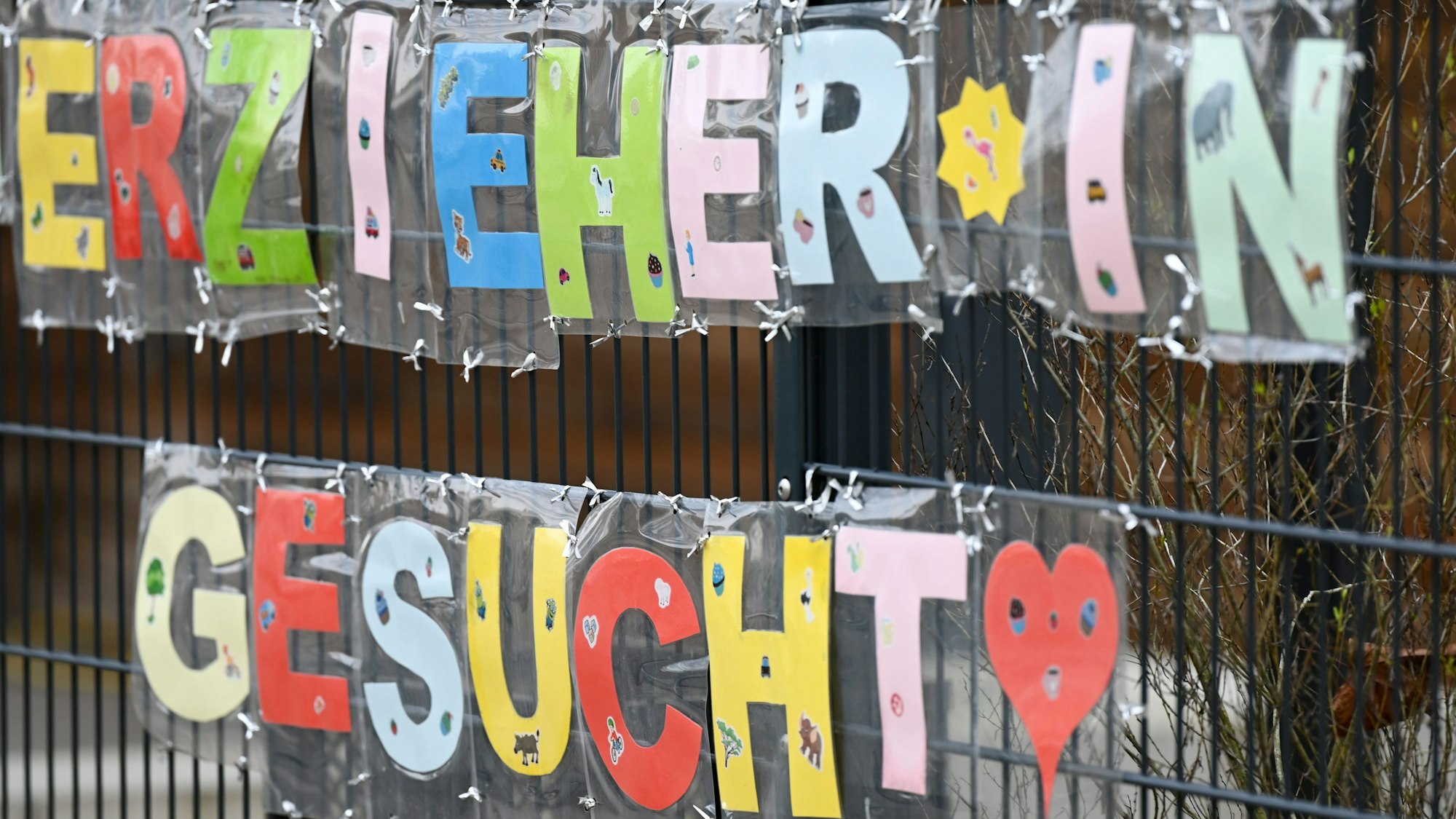 Mit bunten Buchstaben sind die Worte „Erzieher*in gesucht“ geschrieben, die an einem Zaun einer Kindertagesstättebefestigt sind.