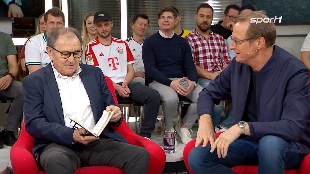 Ewald Lienen schaut beim Sport1-„Doppelpass“ in sein Notizbuch, auf das ihn Florian König gerade angesprochen hat.