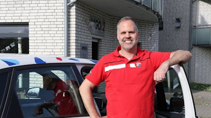 Ein Mann in rotem T-Shirt steht an einer geöffneten Autotür und lächelt in die Kamera.