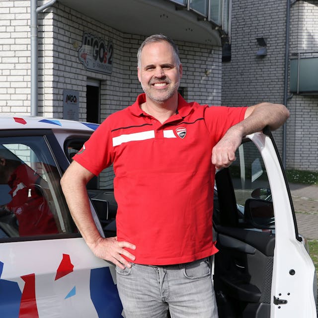 Ein Mann in rotem T-Shirt steht an einer geöffneten Autotür und lächelt in die Kamera.