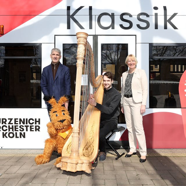 Zu sehen sind drei Menschen, die sich neben einer KVB-Stadtbahn befinden. Einer sitzt an einer Harfe.