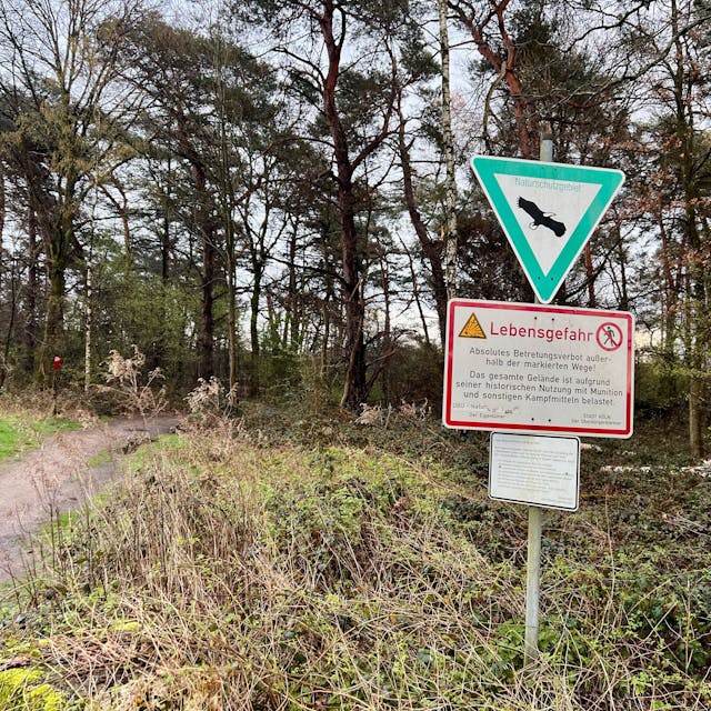 Ein Schild mit dem Hinweis „Lebensgefahr“ in der Wahner Heide weist darauf hin, dass Flächen außerhalb der markierten Wege nicht betreten werden dürfen. Im Hintergrund ist ein Wald zu sehen.