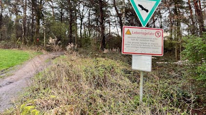 Ein Schild mit dem Hinweis „Lebensgefahr“ in der Wahner Heide weist darauf hin, dass Flächen außerhalb der markierten Wege nicht betreten werden dürfen. Im Hintergrund ist ein Wald zu sehen.