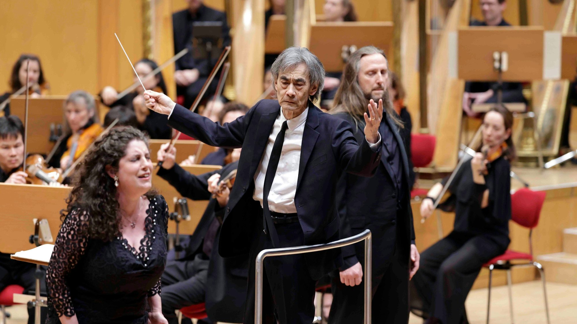 Der dirigent steht auf der Bühne der Philharmonie und breitet die Arme leicht aus, er hält einen Taktstock in de rrechten Hand. An seiner Seite sind die Solisten, im Hintergrund das Orchester.