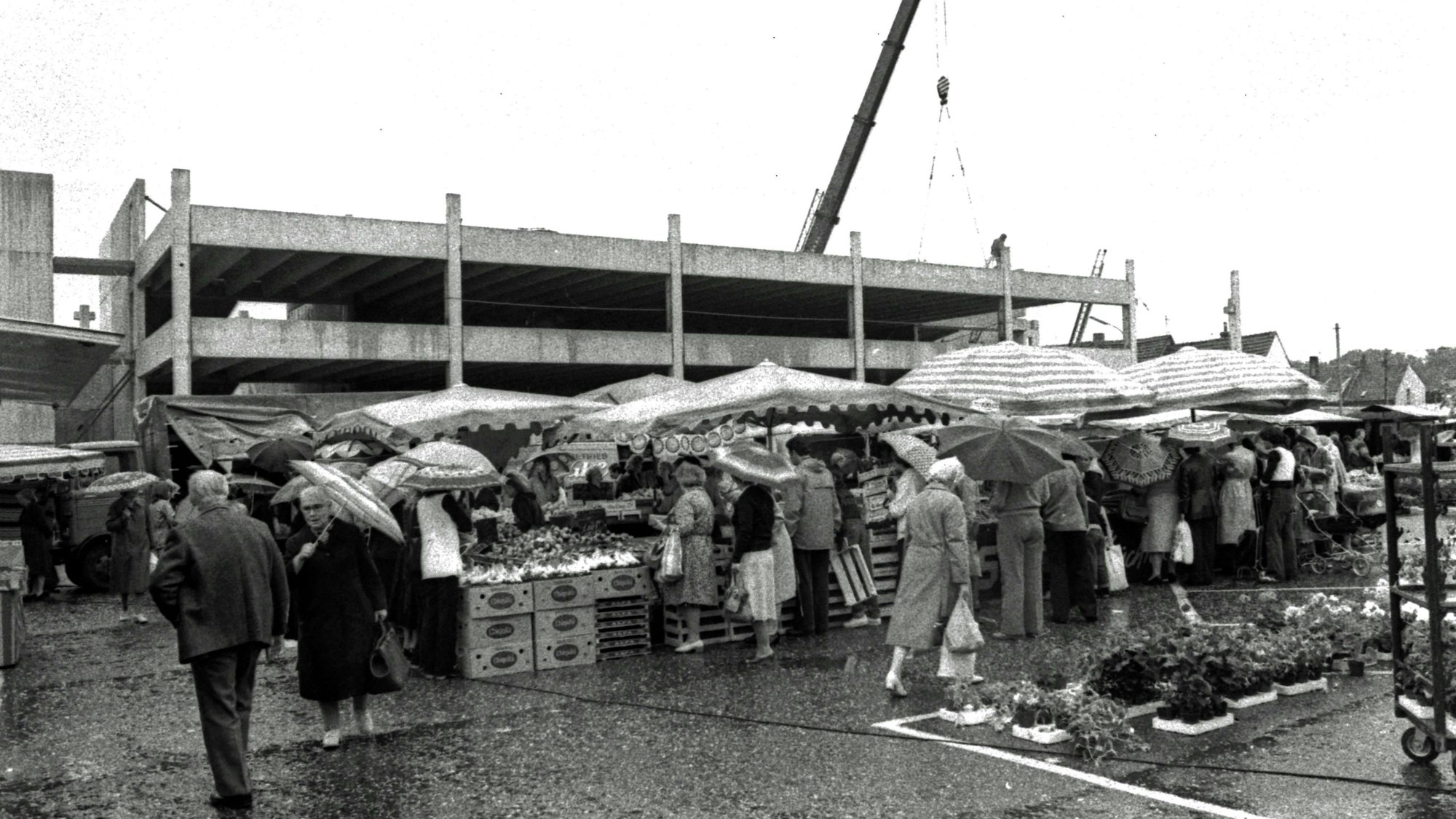 Bei Regenwetter kaufen Menschen an Gemüse- und Blumenständen ein. Im Hintergrund ist der Rohbau des Parkhauses zu sehen.