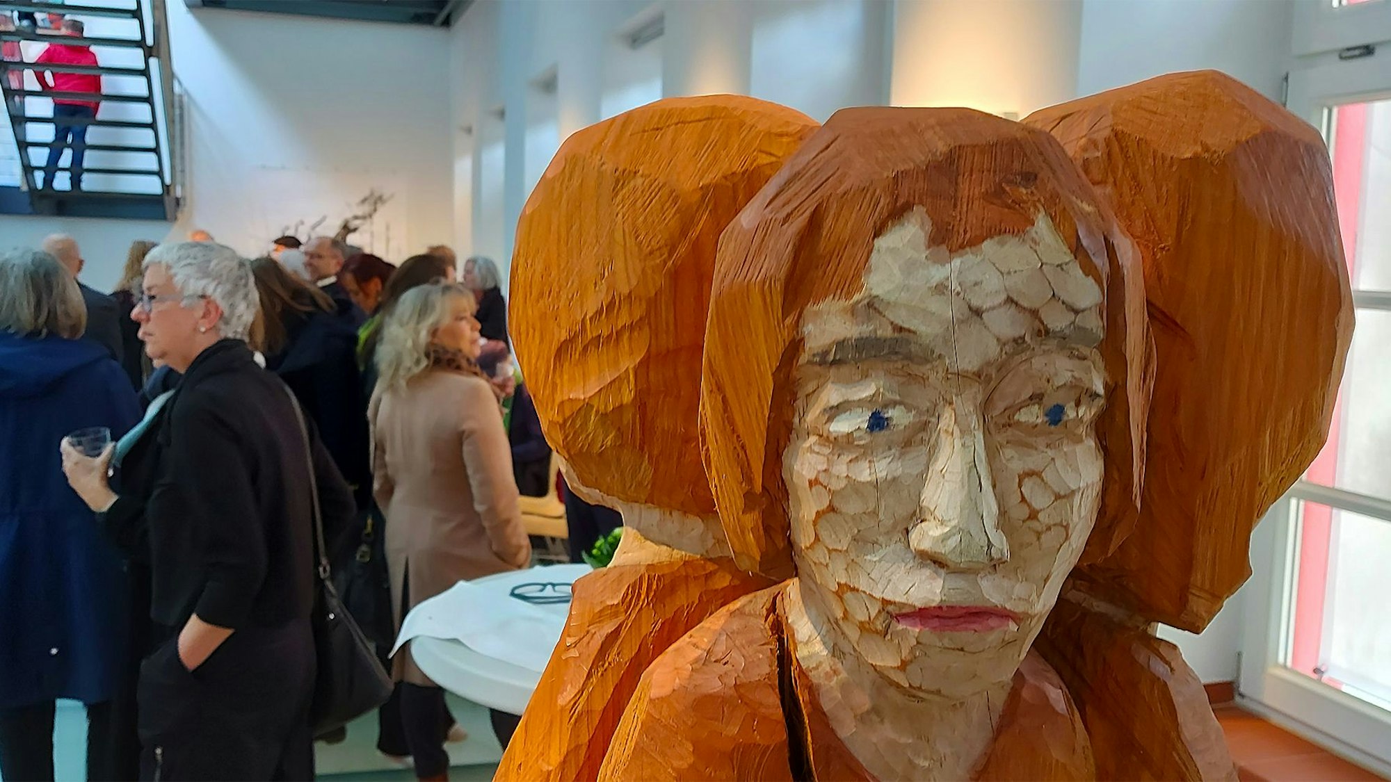 Die Skulptur „Drei Frauen“ von Wolfgang Metzler ist rechts im Bild zu sehen, links im Hintergrund schauen sich Besucher die Ausstellung im Kunstforum Eifel in Gemünd an.