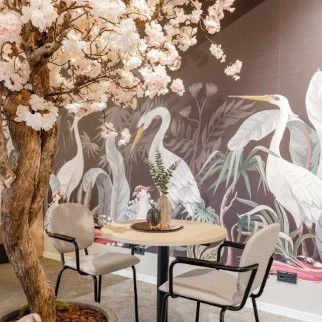 Zu sehen ist ein Tisch in einem Café mit Vogeltapete und einem Baum mit rosafarbenen Blüten.