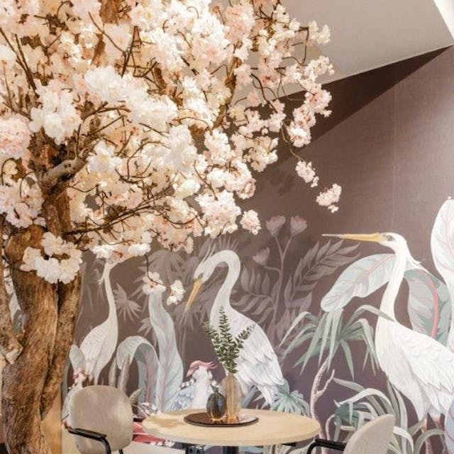Zu sehen ist ein Tisch in einem Café mit Vogeltapete und einem Baum mit rosafarbenen Blüten.
