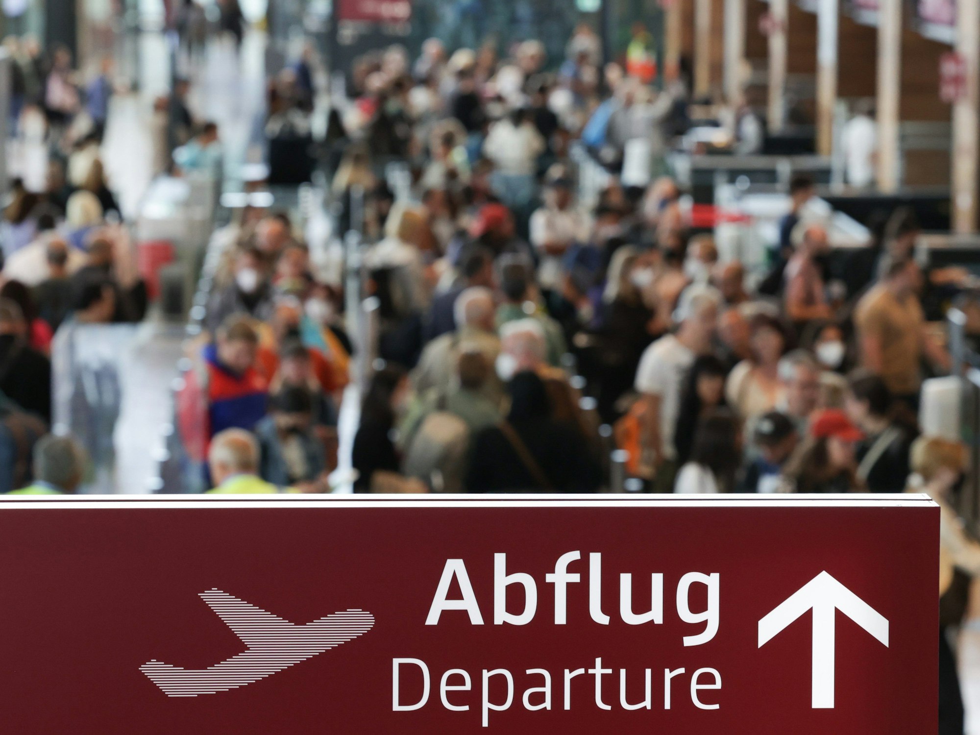 Am Flughafen Berlin Brandenburg BER stehen zahlreiche Reisende in einer Schlange zur Sicherheitskontrolle hinter einem Schild mit der Aufschrift „Abflug“.