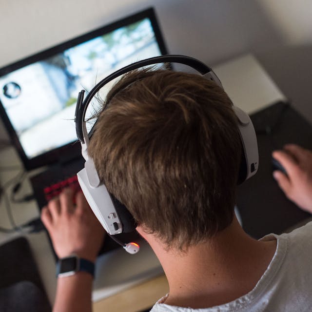 Ein Jugendlicher sitzt  mit einem Headset vor einem Laptop und spielt ein Online-Computerspiel.