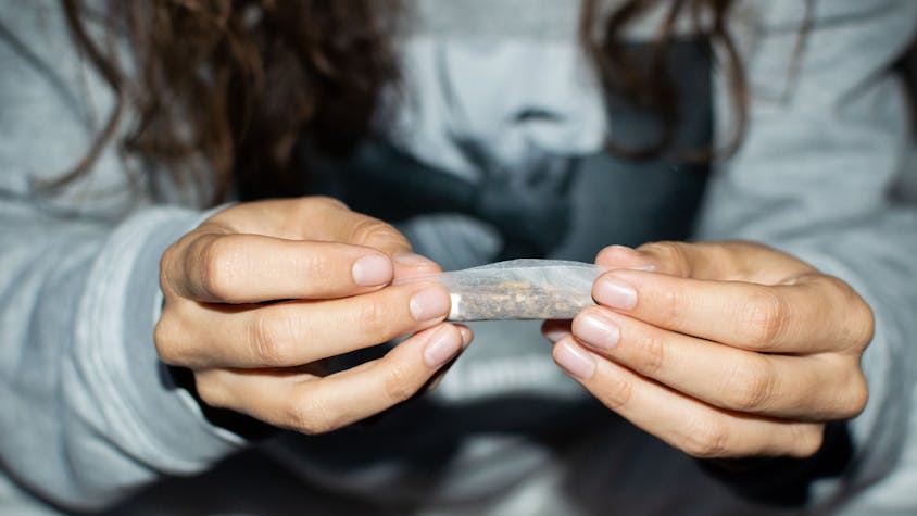 Eine junge Frau rollt nachts einen Marihuana-Joint in einer Straße. Zu sehen sind ihre Hände, in denen sie eine Cannabis-Zigarette hält.  