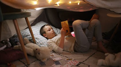 Ein Junge liegt in einer selbstgebauten Höhle mit Sternenhimmel und liest ein Buch.