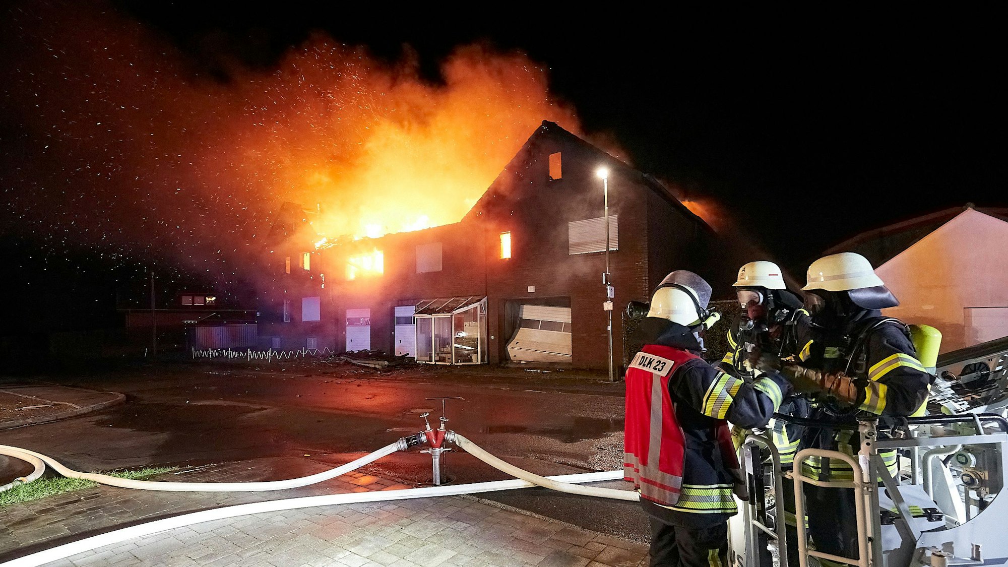 Mehrere Feuerwehrleute stehen vor einem brennenden Haus.